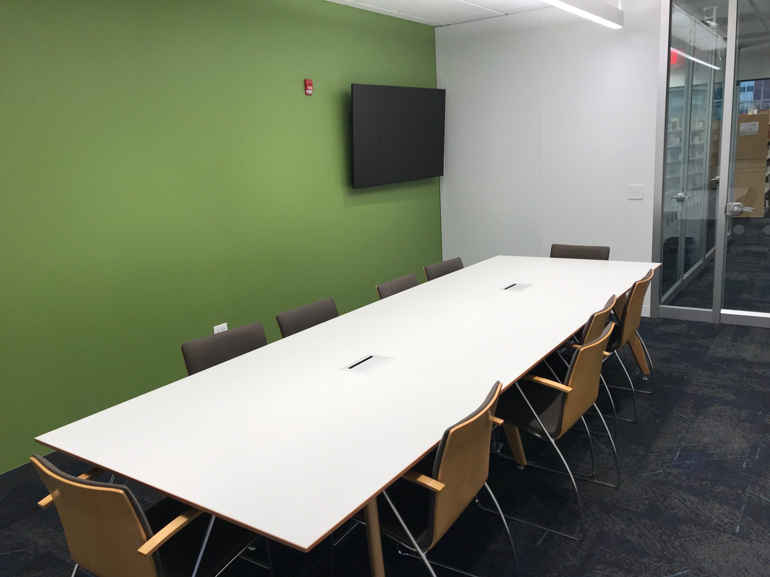 Image of conference room at Aspen Drive Library, shows a long conference table with eight chairs around it. A monitor is in the corner.