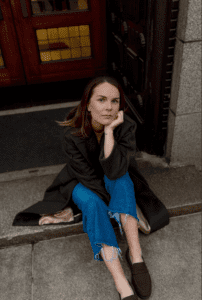 An image of debut author Caro Clare Burke. A white woman with brunette hair, jeans, and a black coat looks up at the camera